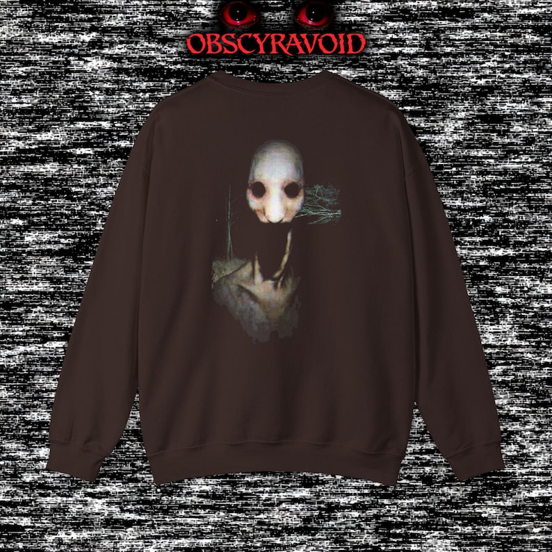 This analog horror sweatshirt features a chilling dual-sided design. The front shows a dark, grainy forest scene with glitchy text reading “I can feel it breathing behind me.” The back displays a disturbing pale creature emerging from the shadows, created with VHS static, noise, and eerie textures. Perfect for horror fans who love unsettling artwork, dark aesthetics, creepy forest themes, and lost-footage style designs.