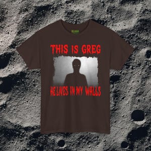 This horror T-shirt features a shadowy, humanoid figure with glowing eyes standing behind a textured, distorted wall pattern. Above it, the phrase “THIS IS GREG” is written in bold, dripping red letters, with “HE LIVES IN MY WALLS” printed below in the same eerie style. The entire design has an analog horror texture, grainy shadows, and unsettling contrast, making it perfect for fans of creepy artwork, scary graphic tees, and surreal dark aesthetics.