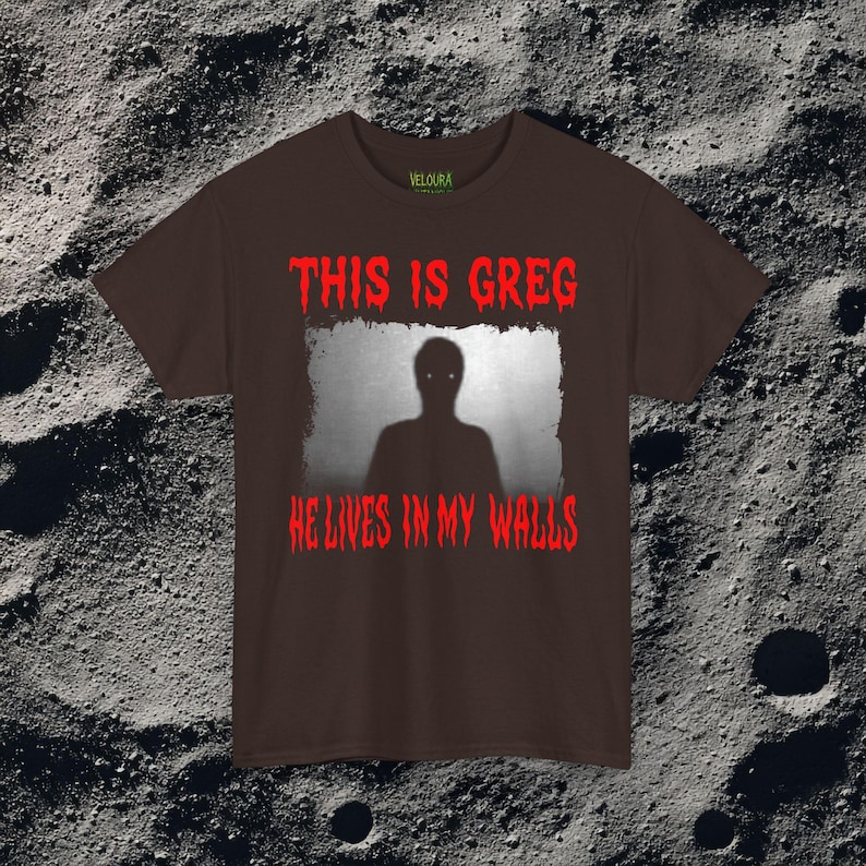 This horror T-shirt features a shadowy, humanoid figure with glowing eyes standing behind a textured, distorted wall pattern. Above it, the phrase “THIS IS GREG” is written in bold, dripping red letters, with “HE LIVES IN MY WALLS” printed below in the same eerie style. The entire design has an analog horror texture, grainy shadows, and unsettling contrast, making it perfect for fans of creepy artwork, scary graphic tees, and surreal dark aesthetics.
