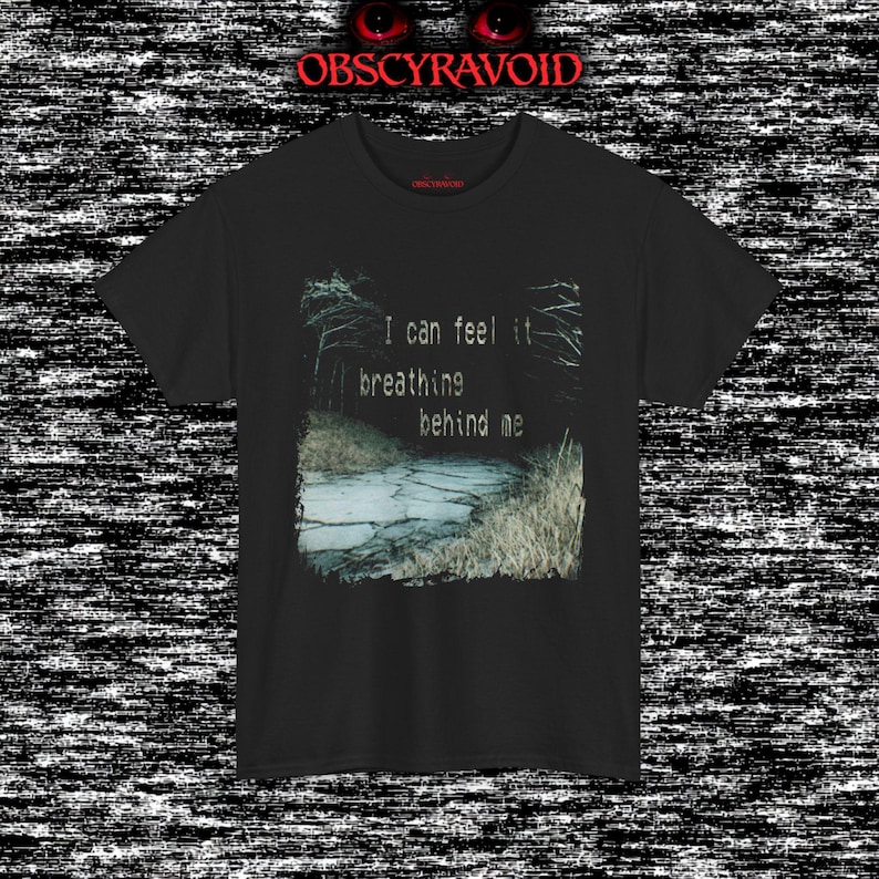 This analog horror T-shirt features a distressed VHS-style forest scene with eerie text overlay, combined with a terrifying pale mask-like creature emerging from darkness. Grainy textures, glitch effects, and cold blue tones create a creepy lost-footage atmosphere. Printed on a soft, high-quality shirt perfect for horror fans, dark aesthetic lovers, and anyone who enjoys unsettling, cinematic artwork.
