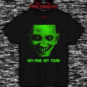 Op de afbeelding: Een zwart T-shirt met korte mouwen en een felgroene, gepixelde afbeelding van een menselijk gezicht met grote ogen en een brede grijns. Onder het gezicht staat de tekst 404 PAGE NOT FOUND in het groen. Bovenaan staat de merknaam OBSCYRAVOID.