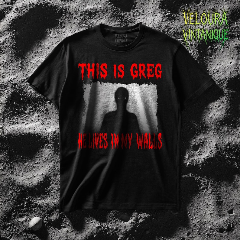 This horror T-shirt features a shadowy, humanoid figure with glowing eyes standing behind a textured, distorted wall pattern. Above it, the phrase “THIS IS GREG” is written in bold, dripping red letters, with “HE LIVES IN MY WALLS” printed below in the same eerie style. The entire design has an analog horror texture, grainy shadows, and unsettling contrast, making it perfect for fans of creepy artwork, scary graphic tees, and surreal dark aesthetics.