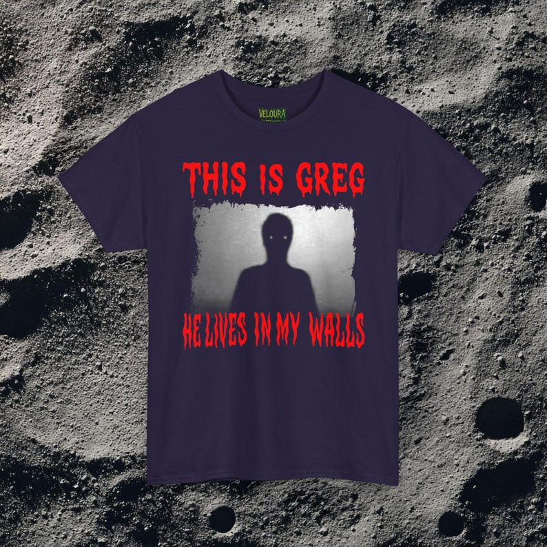 This horror T-shirt features a shadowy, humanoid figure with glowing eyes standing behind a textured, distorted wall pattern. Above it, the phrase “THIS IS GREG” is written in bold, dripping red letters, with “HE LIVES IN MY WALLS” printed below in the same eerie style. The entire design has an analog horror texture, grainy shadows, and unsettling contrast, making it perfect for fans of creepy artwork, scary graphic tees, and surreal dark aesthetics.