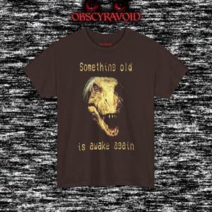 This analog horror–inspired T-shirt features a terrifying decayed dinosaur with human-like eyes and sharp teeth under the phrase “Something old is awake again.” The design combines VHS-style grain, eerie lighting, and vintage texture to create a haunting prehistoric horror vibe. Perfect for analog horror fans, dinosaur lovers, and retro-style collectors. Printed on high-quality fabric for lasting comfort and style.