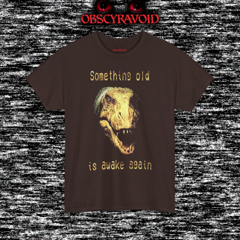 This analog horror–inspired T-shirt features a terrifying decayed dinosaur with human-like eyes and sharp teeth under the phrase “Something old is awake again.” The design combines VHS-style grain, eerie lighting, and vintage texture to create a haunting prehistoric horror vibe. Perfect for analog horror fans, dinosaur lovers, and retro-style collectors. Printed on high-quality fabric for lasting comfort and style.