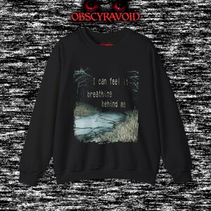 This analog horror sweatshirt features a chilling dual-sided design. The front shows a dark, grainy forest scene with glitchy text reading “I can feel it breathing behind me.” The back displays a disturbing pale creature emerging from the shadows, created with VHS static, noise, and eerie textures. Perfect for horror fans who love unsettling artwork, dark aesthetics, creepy forest themes, and lost-footage style designs.