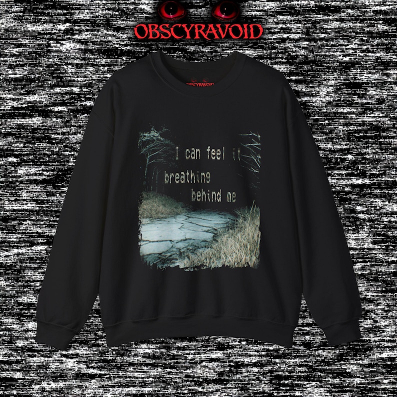 This analog horror sweatshirt features a chilling dual-sided design. The front shows a dark, grainy forest scene with glitchy text reading “I can feel it breathing behind me.” The back displays a disturbing pale creature emerging from the shadows, created with VHS static, noise, and eerie textures. Perfect for horror fans who love unsettling artwork, dark aesthetics, creepy forest themes, and lost-footage style designs.