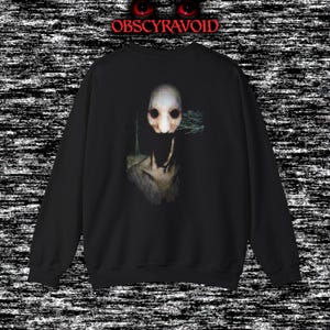 This analog horror sweatshirt features a chilling dual-sided design. The front shows a dark, grainy forest scene with glitchy text reading “I can feel it breathing behind me.” The back displays a disturbing pale creature emerging from the shadows, created with VHS static, noise, and eerie textures. Perfect for horror fans who love unsettling artwork, dark aesthetics, creepy forest themes, and lost-footage style designs.