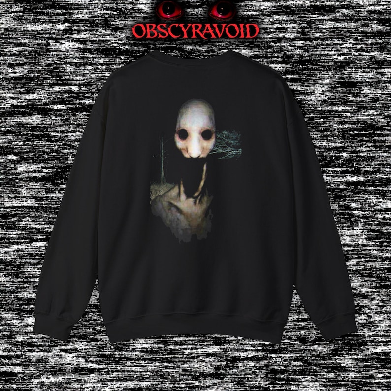 This analog horror sweatshirt features a chilling dual-sided design. The front shows a dark, grainy forest scene with glitchy text reading “I can feel it breathing behind me.” The back displays a disturbing pale creature emerging from the shadows, created with VHS static, noise, and eerie textures. Perfect for horror fans who love unsettling artwork, dark aesthetics, creepy forest themes, and lost-footage style designs.