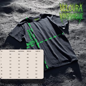 May include: Dark gray t-shirt with the text "VELOURA VINTANIQUE" in green, along with a size chart showing width and length in inches and centimeters. The shirt has a crew neck and short sleeves. The image is on a textured surface.
