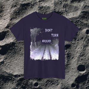 This analog horror-inspired T-shirt features the phrase “DON’T TURN AROUND” printed across a dark, eerie forest road under moonlight. On the other side, a creepy, distorted bunny character stares with a wide unsettling grin. The design blends glitchcore, VHS static, and nightmare-style textures for a perfect horror aesthetic. Printed on soft, high-quality fabric, ideal for horror fans and lovers of dark surreal art.