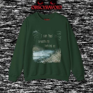 This analog horror sweatshirt features a chilling dual-sided design. The front shows a dark, grainy forest scene with glitchy text reading “I can feel it breathing behind me.” The back displays a disturbing pale creature emerging from the shadows, created with VHS static, noise, and eerie textures. Perfect for horror fans who love unsettling artwork, dark aesthetics, creepy forest themes, and lost-footage style designs.