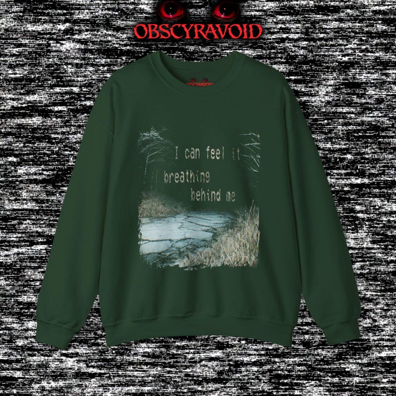 This analog horror sweatshirt features a chilling dual-sided design. The front shows a dark, grainy forest scene with glitchy text reading “I can feel it breathing behind me.” The back displays a disturbing pale creature emerging from the shadows, created with VHS static, noise, and eerie textures. Perfect for horror fans who love unsettling artwork, dark aesthetics, creepy forest themes, and lost-footage style designs.