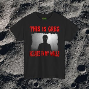 This horror T-shirt features a shadowy, humanoid figure with glowing eyes standing behind a textured, distorted wall pattern. Above it, the phrase “THIS IS GREG” is written in bold, dripping red letters, with “HE LIVES IN MY WALLS” printed below in the same eerie style. The entire design has an analog horror texture, grainy shadows, and unsettling contrast, making it perfect for fans of creepy artwork, scary graphic tees, and surreal dark aesthetics.