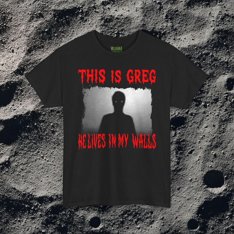 This horror T-shirt features a shadowy, humanoid figure with glowing eyes standing behind a textured, distorted wall pattern. Above it, the phrase “THIS IS GREG” is written in bold, dripping red letters, with “HE LIVES IN MY WALLS” printed below in the same eerie style. The entire design has an analog horror texture, grainy shadows, and unsettling contrast, making it perfect for fans of creepy artwork, scary graphic tees, and surreal dark aesthetics.