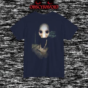 This analog horror T-shirt features a distressed VHS-style forest scene with eerie text overlay, combined with a terrifying pale mask-like creature emerging from darkness. Grainy textures, glitch effects, and cold blue tones create a creepy lost-footage atmosphere. Printed on a soft, high-quality shirt perfect for horror fans, dark aesthetic lovers, and anyone who enjoys unsettling, cinematic artwork.