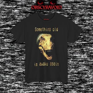 This analog horror–inspired T-shirt features a terrifying decayed dinosaur with human-like eyes and sharp teeth under the phrase “Something old is awake again.” The design combines VHS-style grain, eerie lighting, and vintage texture to create a haunting prehistoric horror vibe. Perfect for analog horror fans, dinosaur lovers, and retro-style collectors. Printed on high-quality fabric for lasting comfort and style.