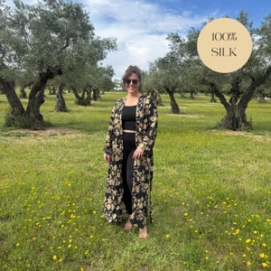 May include: A black kimono with a gold floral pattern. The kimono is made of silk and has a long, flowing design. The text "100% SILK" is in a circle in the upper right corner. The person is standing in a field of grass and yellow flowers.