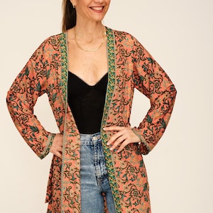 Könnte beinhalten: Lange, offen geschnittene Kimono-Jacke mit floralem Muster in Koralle, Grün und Schwarz. Die Jacke hat grüne Verzierungen und wird über einem schwarzen Oberteil und einer blauen Jeans getragen. Eine goldene Halskette ist ebenfalls sichtbar.