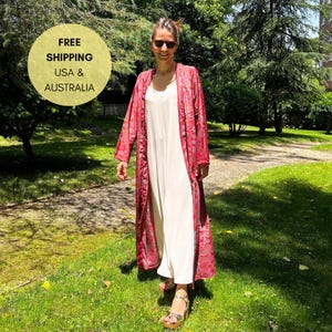 May include: A woman wearing a long, open red patterned robe over a white dress. The robe has a floral design. She is wearing sunglasses and sandals, standing on a grassy area with trees in the background.