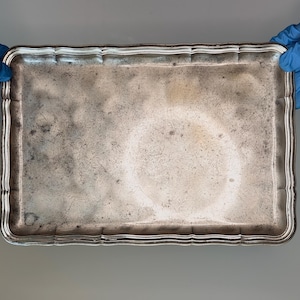 Puede incluir: Una bandeja rectangular de metal plateado con un borde festoneado y una superficie central lisa y plana. La bandeja presenta un acabado desgastado y deslustrado con arañazos visibles y manchas de pátina oscura en toda la base.