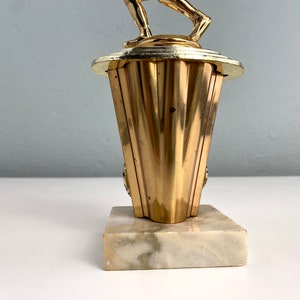 May include: Gold-colored trophy with a figure of a person holding a ball on top. The base is made of marble with a white and gray pattern.