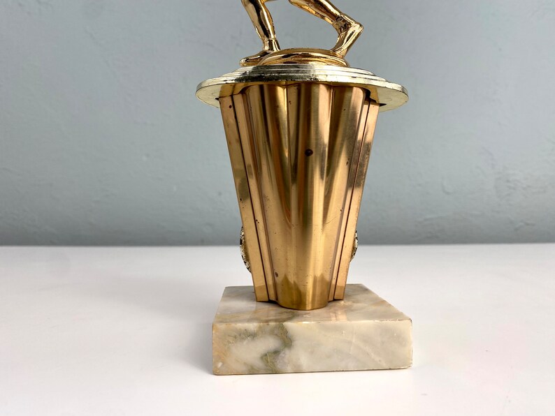 May include: Gold-colored trophy with a figure of a person holding a ball on top. The base is made of marble with a white and gray pattern.