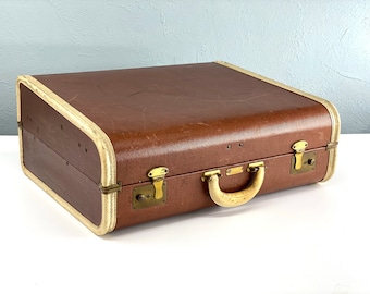 May include: A vintage brown suitcase with a cream-colored trim and brass hardware. The suitcase has a rounded top and a handle with a cream-colored grip.