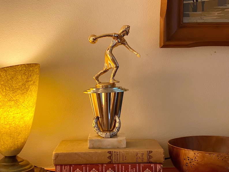 May include: A gold-toned trophy featuring a female figure holding a bowling ball. The trophy is on a marble base with a gold-toned decorative band.