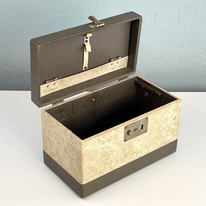 May include: An open vintage metal box with a brown exterior and a cream-colored interior. The box has a lock and a metal latch.