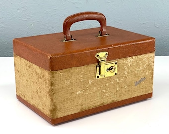 May include: A vintage brown leather and tan fabric travel case with a brass lock and handle. The case has a "Tweedie" brand name on the front.