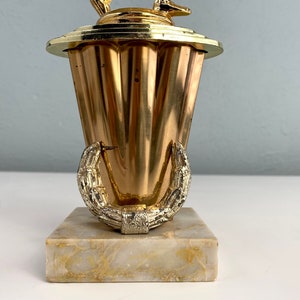 May include: A gold-colored trophy with a stylized deer figure on top. The trophy has a fluted base and a laurel wreath design around the bottom. The base is made of marble.