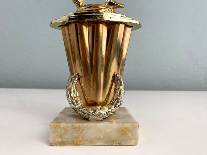 May include: A gold-colored trophy with a stylized deer figure on top. The trophy has a fluted base and a laurel wreath design around the bottom. The base is made of marble.