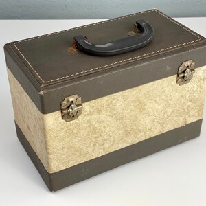 May include: A vintage brown and cream cardboard box with a black plastic handle and metal clasps. The box has a textured surface and a stitched seam around the top edge.