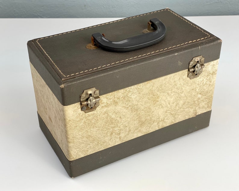 May include: A vintage brown and cream cardboard box with a black plastic handle and metal clasps. The box has a textured surface and a stitched seam around the top edge.