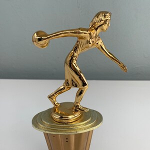 May include: Gold-colored trophy of a person bowling with a ball in their hand. The trophy is on a round base with a textured surface.