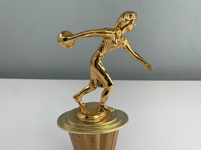 May include: Gold-colored trophy of a person bowling with a ball in their hand. The trophy is on a round base with a textured surface.