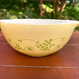May include: A vintage yellow mixing bowl with a white interior and a floral design. The bowl has a light yellow exterior with a green floral pattern. It features two handles and is suitable for kitchen use.