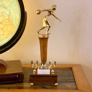 May include: A gold-toned trophy with a female figure holding a bowling ball. The trophy has a wooden base with a marble top and two gold wings on the sides.