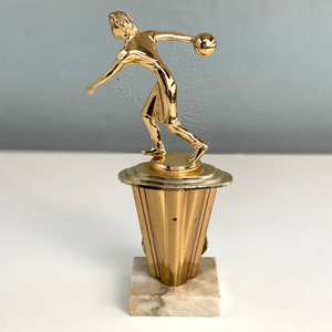 May include: Gold-toned bowling trophy featuring a figurine of a person throwing a bowling ball. The trophy has a marble base.