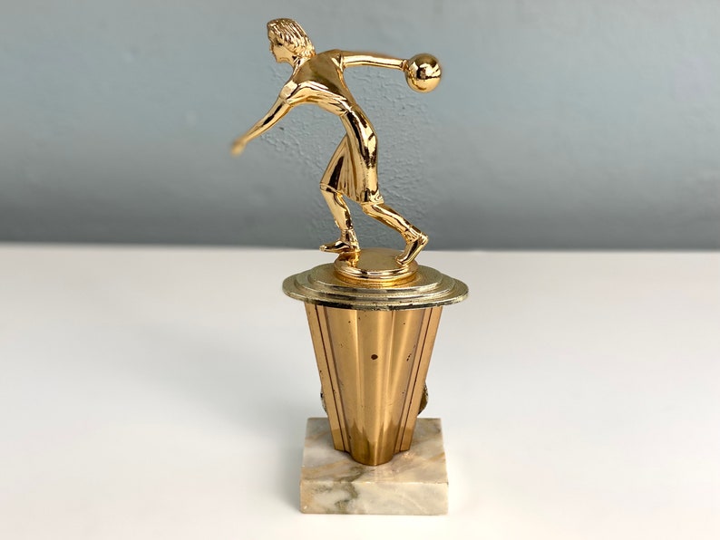 May include: Gold-toned bowling trophy featuring a figurine of a person throwing a bowling ball. The trophy has a marble base.