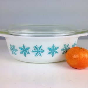 May include: White oval casserole dish with a clear glass lid. The dish has a turquoise snowflake design around the rim. A small orange sits in front of the dish.