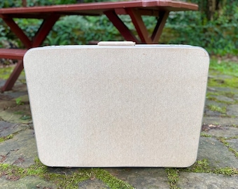 May include: A vintage beige suitcase with a textured surface. The suitcase has a rectangular shape with rounded corners and a silver trim. It features a handle on top and is set against a natural outdoor backdrop.
