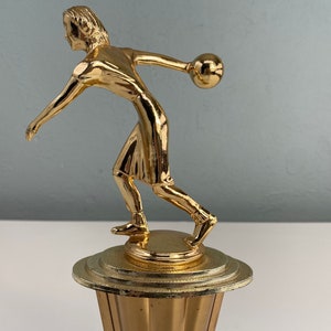 May include: Gold-colored trophy of a person bowling. The person is in mid-throw, with a bowling ball in their hand. The trophy is on a round base with a fluted design.