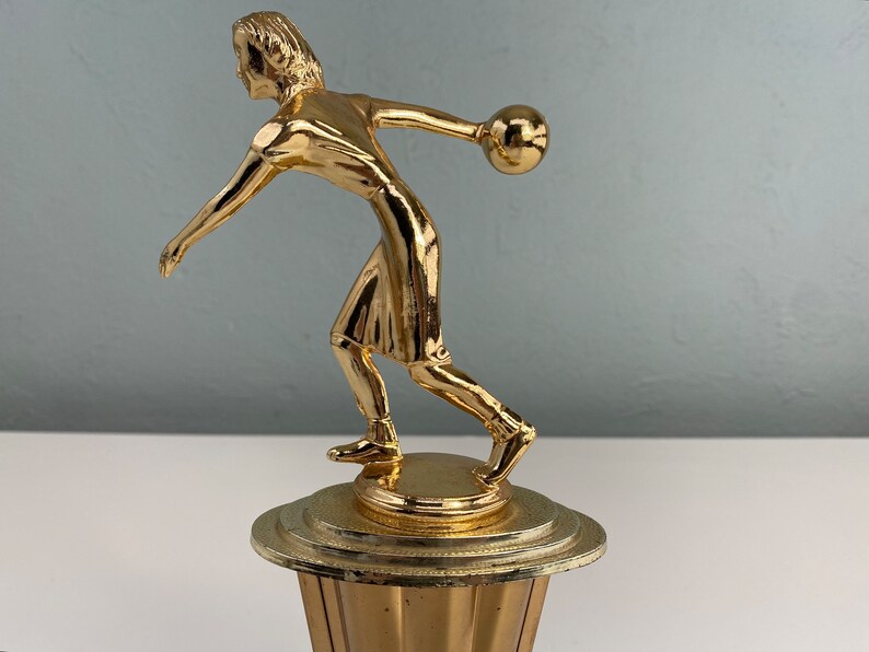 May include: Gold-colored trophy of a person bowling. The person is in mid-throw, with a bowling ball in their hand. The trophy is on a round base with a fluted design.