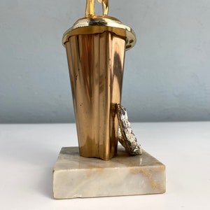 May include: Gold-colored trophy with a figure of a person holding a ball on top. The base is made of white marble.