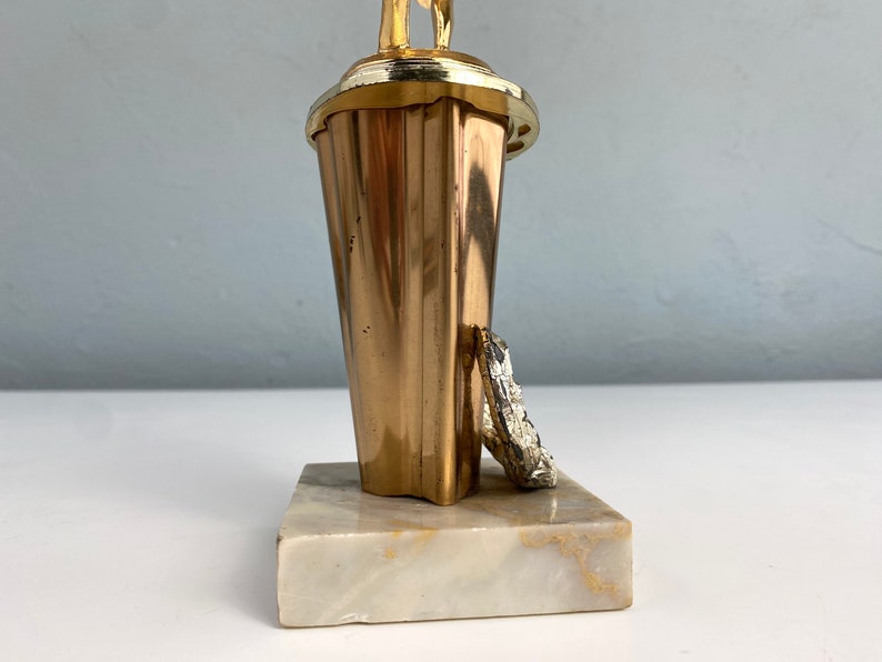 May include: Gold-colored trophy with a figure of a person holding a ball on top. The base is made of white marble.