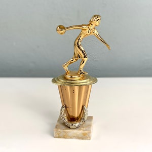 May include: Gold-colored trophy with a female figure holding a bowling ball. The base is a gold-colored pedestal with a marble-like base.