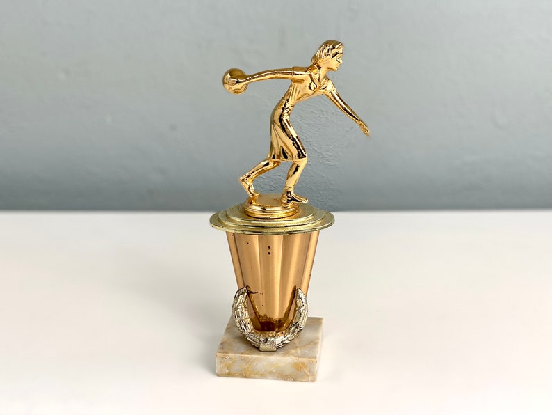 May include: Gold-colored trophy with a female figure holding a bowling ball. The base is a gold-colored pedestal with a marble-like base.