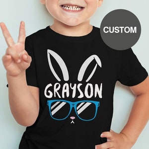 May include: Black t-shirt with a white bunny rabbit graphic wearing blue sunglasses and the name "GRAYSON" printed on the front. The shirt is being worn by a young child.